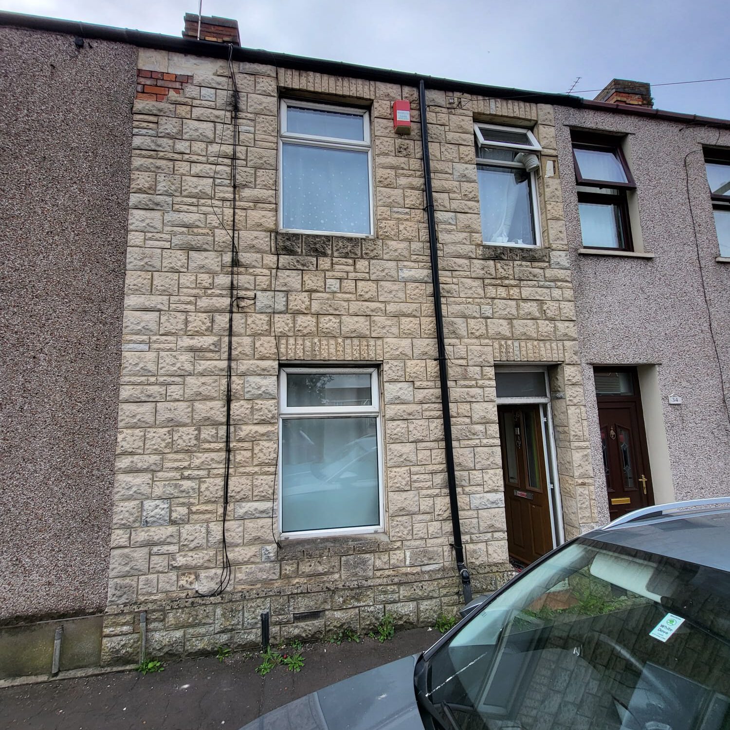 A two-story stone terraced house with a large window on the ground floor and a smaller window on the first floor. The building has a brown door to the right of the ground floor window and a brick chimney. A gray car is parked in front of the house.